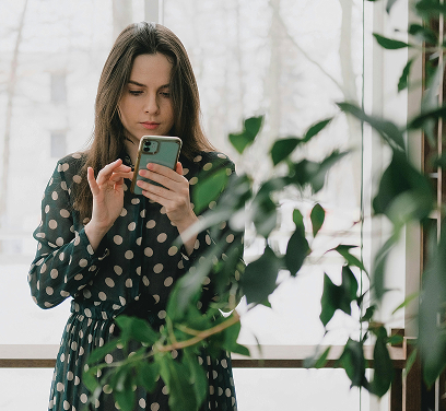 A woman with long, dark hair, wearing a dark polka-dotted dress, is looking at and interacting with her smartphone. She is standing indoors near a window, with some green foliage in the foreground on the right side of the image.