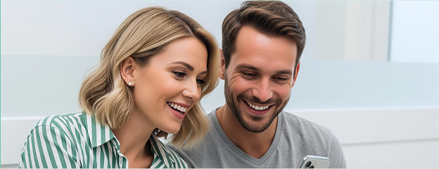 A man and a woman are looking down and smiling, likely at a smartphone held by the man, which is partially visible at the bottom right. The woman, on the left, has blonde hair and is wearing a green and white striped shirt. The man, on the right, has dark hair and is wearing a grey t-shirt. They both appear happy and engaged.