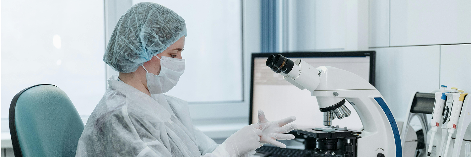 A person wearing a lab coat, hairnet, and face mask is seated at a desk, looking towards a microscope. Their hands are gloved, and one hand is positioned near the microscope. In the background, a computer monitor is visible, and other lab equipment can be seen on the right side of the desk.